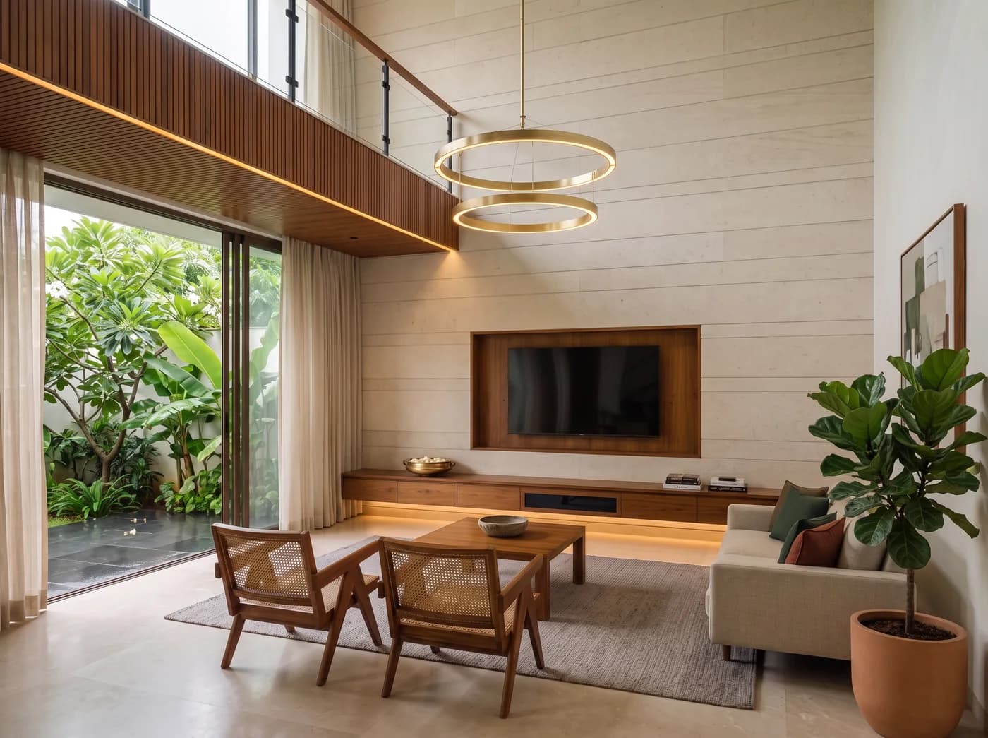Double-height villa living room on a monsoon afternoon: a wall of honed limestone in coursed bands with a teak-lined television niche and a long floating teak console lit from beneath, a brass urli of frangipani on it, full-height sliding glass onto a courtyard of frangipani and banana leaf over wet kota, sheer handloom curtains, a teak-slat bulkhead with a light line under a glass-and-teak mezzanine rail, a two-ring brass chandelier on a long rod, an oatmeal handloom sofa with green and rust cushions, a pair of teak-and-cane Chandigarh chairs, a solid teak coffee table on a warm-grey rug, a large abstract in a teak frame and a fiddle-leaf fig on a plaster wall, pale honed stone underfoot