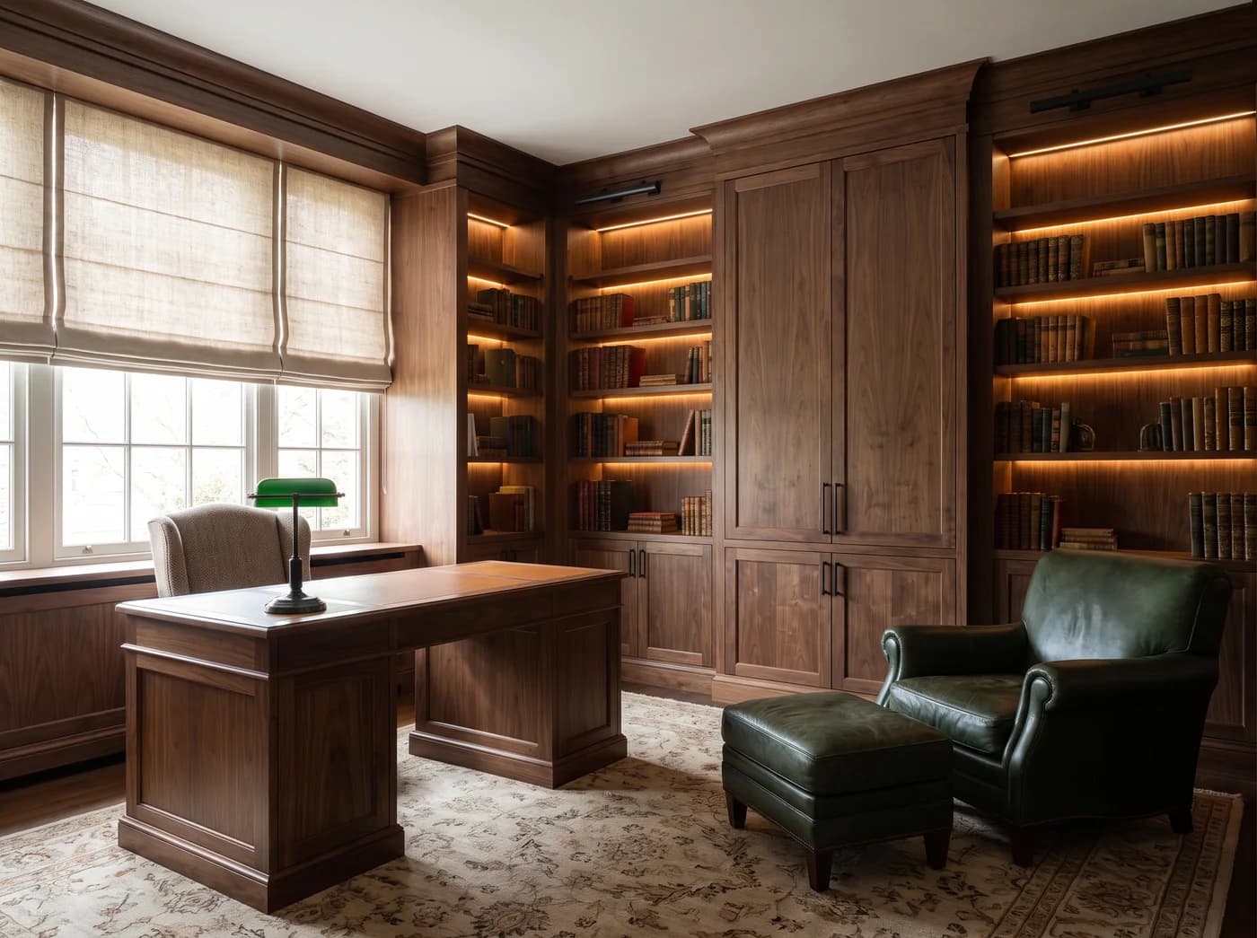 Walnut-panelled study with lit bookshelves on two walls around a central cupboard, black picture lights on the cornice, a leather-topped walnut pedestal desk in front of a linen roman blind with a green banker's lamp, a small upholstered desk chair, a green leather club chair and ottoman on a hand-knotted rug over a dark wood floor